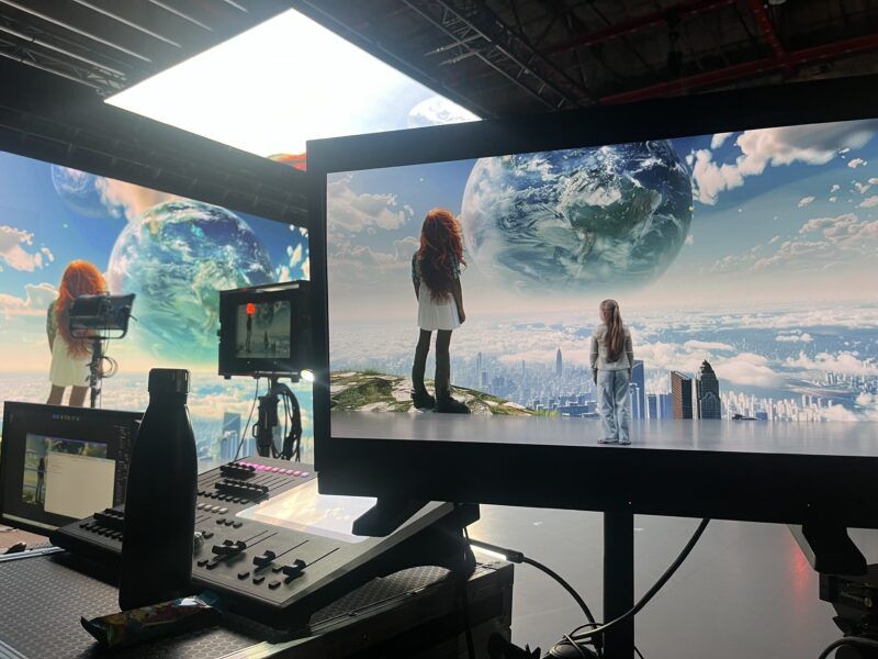Film set with two girls facing a screen showing a surreal city skyline and a giant floating Earth in the sky.
